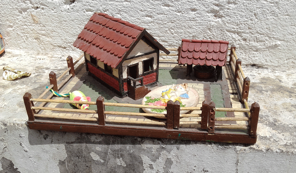 Uma casinha de brinquedo ao lado de um poço de brinquedo com telhado. Um ratinho colorido e um anjinho também estão no conjunto, que é circulado por uma cerca baixa.