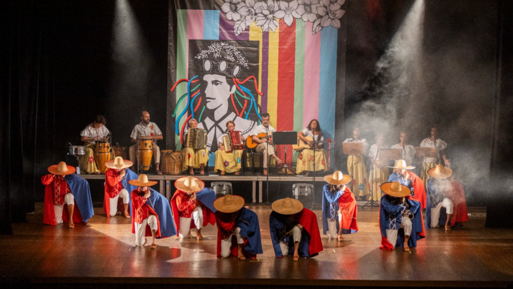 Foto de uma apresentação artística em um palco de teatro. Na parte da frente, dançarinos ajoelhados formam duas fileiras, usando chapéus de palha largos, capas vermelhas e azuis e roupas brancas. Ao fundo, músicos sentados tocam instrumentos como acordeom, violão e tambores. Atrás deles há um grande painel colorido com faixas verticais e o desenho em preto e branco de uma pessoa com flores na cabeça. Dois refletores iluminam o palco de cima, enquanto fumaça cênica cria um efeito dramático ao redor dos artistas.
