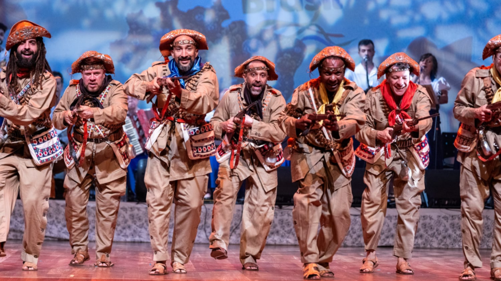 O ambiente é um palco de teatro com piso de madeira clara e um fundo cênico em tons de azul e branco. Em destaque, sete homens avançam em direção ao espectador em uma performance coreografada, todos vestidos com trajes de cangaceiros. Eles usam túnicas e calças de brim em tom cáqui, adornadas com bandoleiras cruzadas no peito, bolsas laterais bordadas e chapéus de couro de aba dobrada com detalhes em relevo. Alguns homens usam lenços coloridos no pescoço em tons de azul, vermelho e amarelo. Com expressões faciais intensas e sorridentes, eles empunham réplicas de fuzis de madeira. Ao fundo, de forma levemente desfocada, aparecem músicos e outros integrantes da companhia.