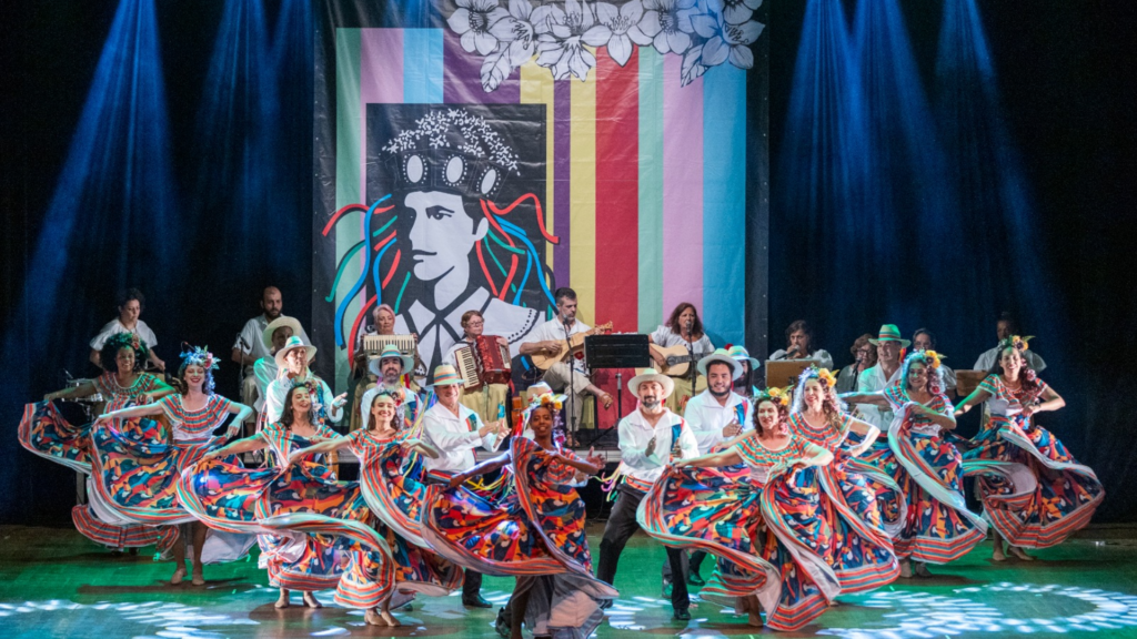 Em um palco de teatro iluminado por refletores, um grupo de dançarinos se apresentam em formação. Eles usam chapéus de aba larga, capas vermelhas e azuis e roupas claras. Ao fundo, músicos tocam instrumentos como acordeom, violão e percussão, sentados em uma plataforma elevada. Atrás deles há um grande painel colorido com listras verticais em tons vibrantes e a ilustração em preto e branco de uma pessoa usando adornos florais na cabeça. Uma leve fumaça cênica espalha-se pelo palco, criando atmosfera dramática para a apresentação cultural.