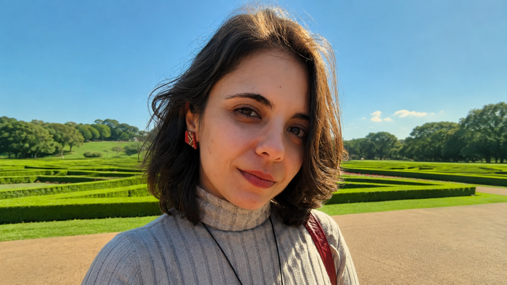 O ambiente é ensolarado e ao ar livre, com um céu azul limpo e luz solar direta que ilumina o lado esquerdo do rosto da mulher. Em primeiro plano, uma jovem de pele clara, olhos castanhos e cabelos castanhos curtos, na altura do queixo, olha diretamente para a câmera com um leve sorriso. Ela veste uma blusa de gola alta em malha canelada cinza claro e usa um brinco geométrico artesanal com detalhes em vermelho e madeira. Ao fundo, destaca-se um jardim de estilo clássico com gramados verdes e arbustos podados em linhas geométricas.