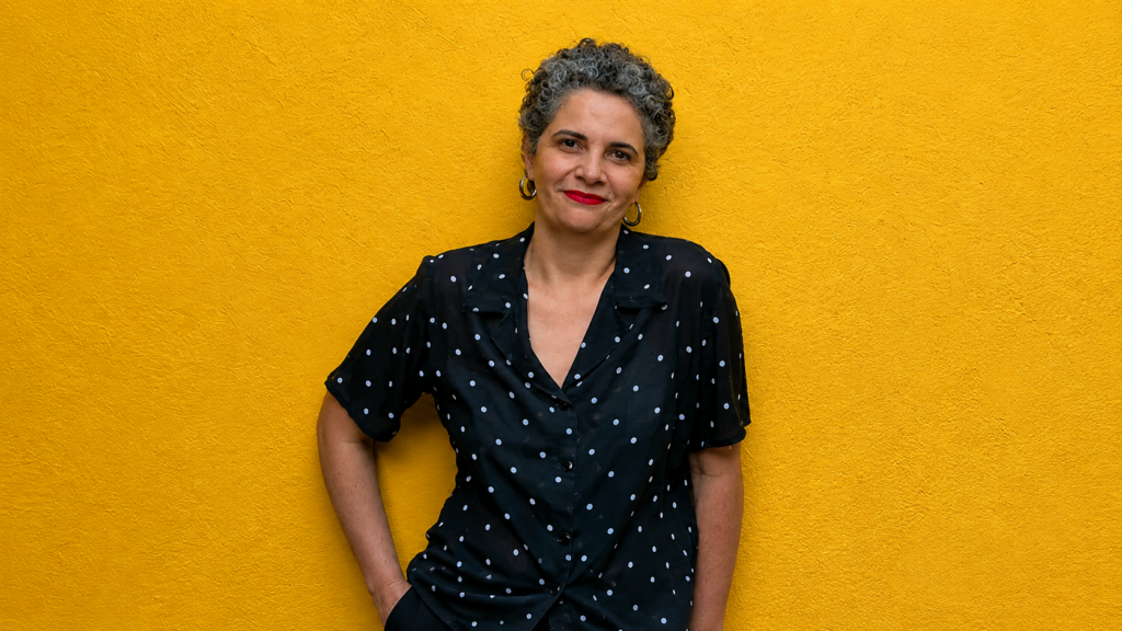 O ambiente é vibrante e minimalista, composto por um fundo de parede com textura granulada em um tom de amarelo intenso. Em plano médio e centralizada, uma mulher de pele clara e expressão serena posa para o retrato. Ela tem cabelos curtos, cacheados e grisalhos, e usa um batom vermelho marcante que contrasta com o fundo. Sua vestimenta consiste em uma camisa de manga curta preta com estampa de poá e gola colarinho. Como acessório, ela utiliza brincos de argola prateados de tamanho médio. A iluminação é frontal e uniforme, destacando as cores e as texturas da cena.
