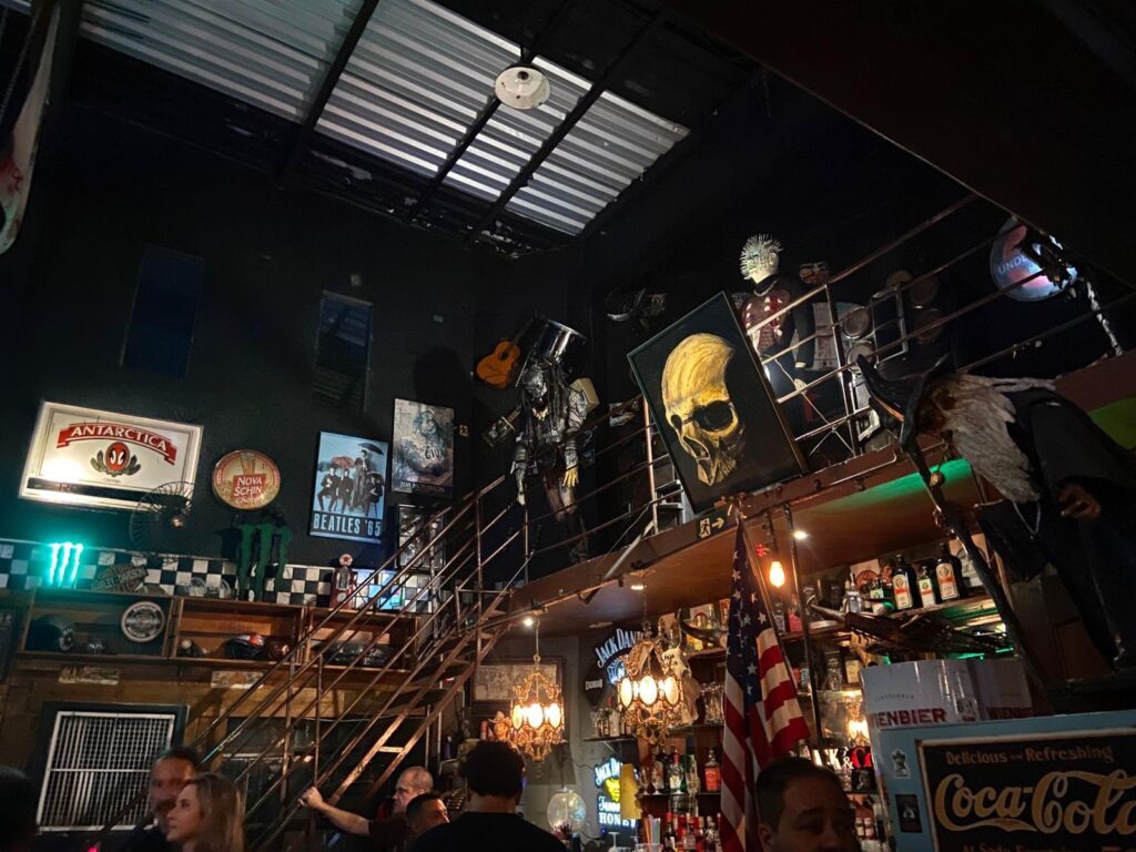 Foto de um interior de uma casa dedicada ao universo do rock. O ambiente é escuro, com iluminação baixa e atmosfera que remete à estética do rock clássico. Ao fundo, uma escada metálica leva a um mezanino decorado com objetos como guitarras penduradas, um grande quadro com a imagem de uma caveira e esculturas de personagens cinematográficos. Nas paredes, placas vintage e pôsteres de marcas de bebidas e bandas do gênero compõem a decoração do espaço. Na parte inferior da imagem, algumas pessoas aparecem reunidas próximas ao balcão, entre garrafas, luminárias, uma bandeira dos Estados Unidos e a escultura de um mago, reforçando o estilo alternativo do espaço.